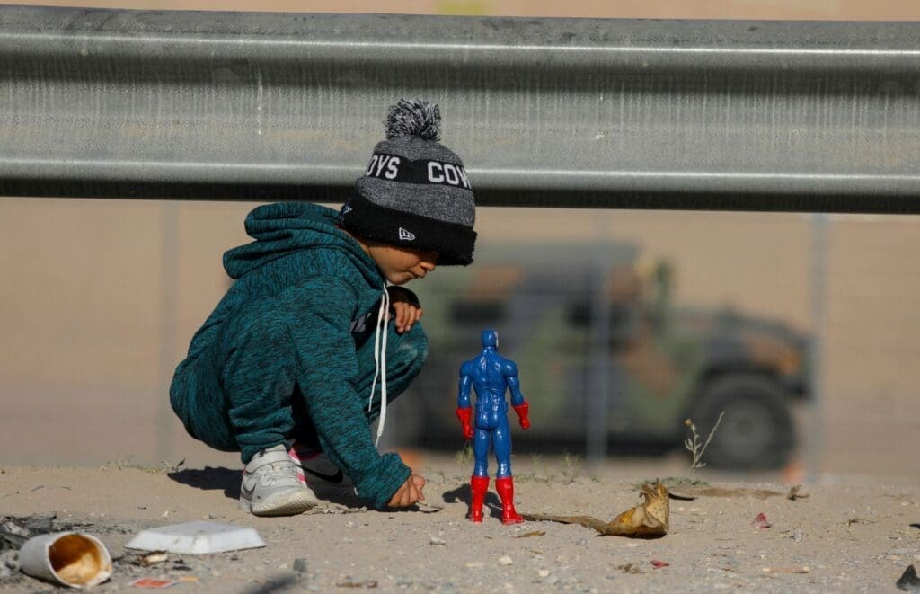 Migrant child crouches down to play with an action figure along the U.S.-Mexico border.