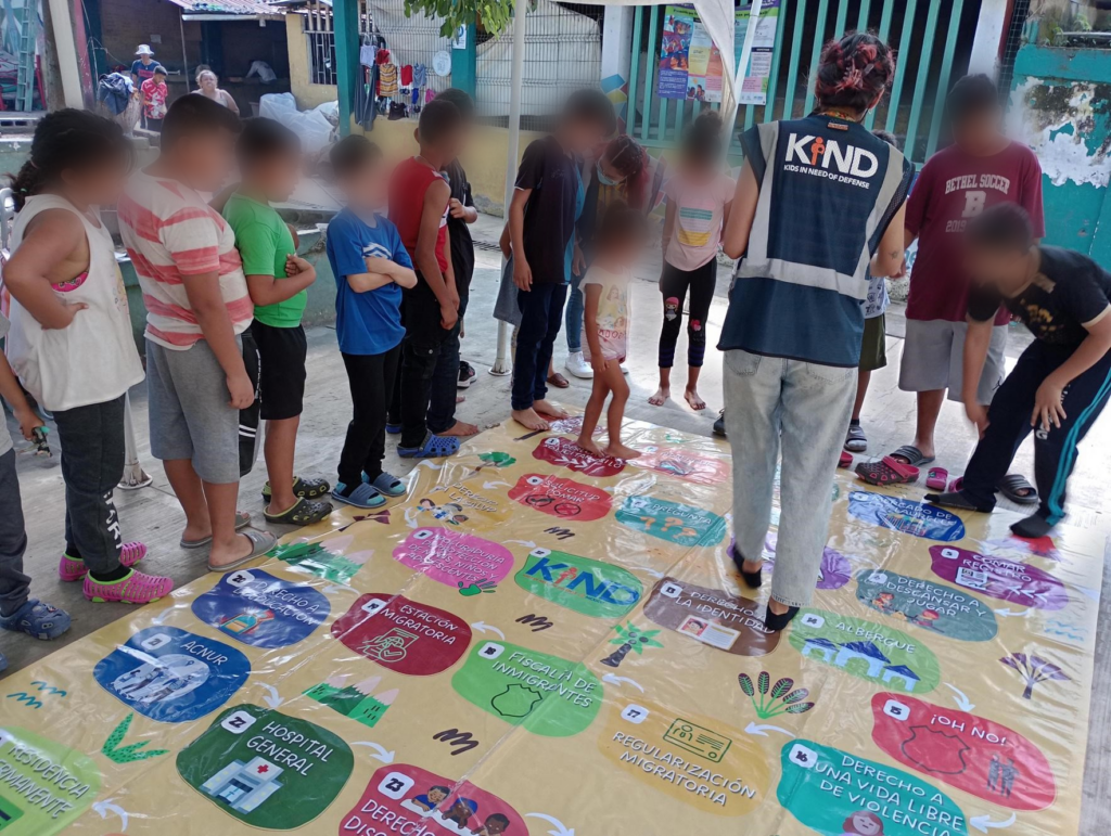 KIND Staff in navy and gray vest talking to a group of children as they stand on a colorful-board game.
