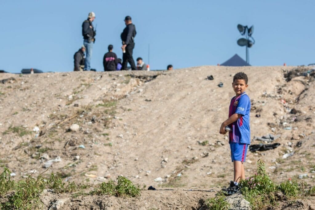 An unaccompanied boy in a blue outfit stands near a rocky hill, staring at the camera.