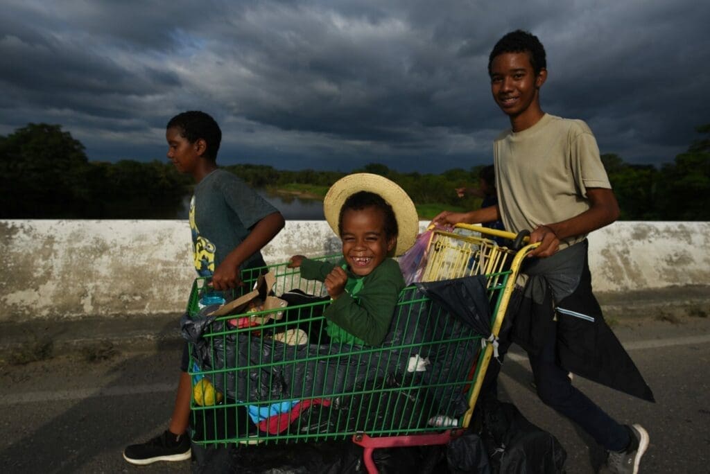 Migrant children laugh and get ahead with the help of a shopping cart with numerous migrants walking towards the US border.