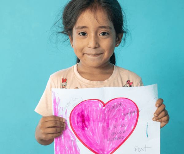 Young girl smiling as she holds up her drawing of her pink heart.