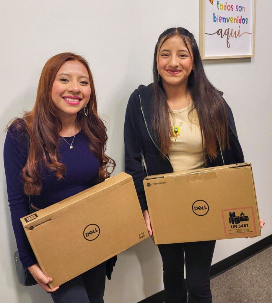 Two young girls with laptops given to them by the Hispanic Heritage Foundation smile at the camera.