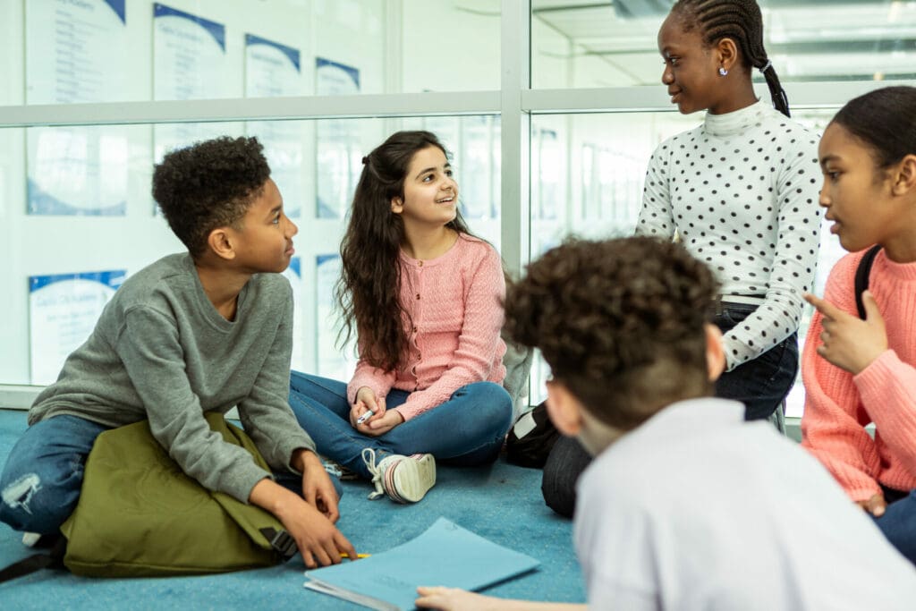 A multiracial group of adolescent boys and girls sit on the floor in the school library and talk while working on a school assignment after class.